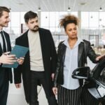 An interracial couple consults with a salesman at a car dealership, exploring vehicle options.