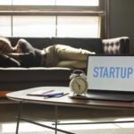 A woman lies on a sofa while a laptop displays 'STARTUP,' capturing a workspace vibe.